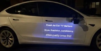 White Tesla at night with a door projection reading Fresh As Can V Barbers, Style. Precision. Consistency. Where quality comes first!