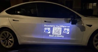 White Tesla at night with a door projection reading Experience the Difference, a QR code and Book on Booksy