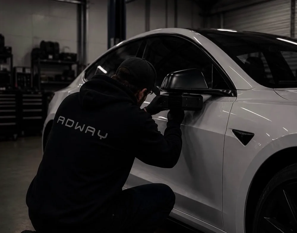 Technician in an Adway hoodie fitting a projector beside the side mirror of a white Tesla