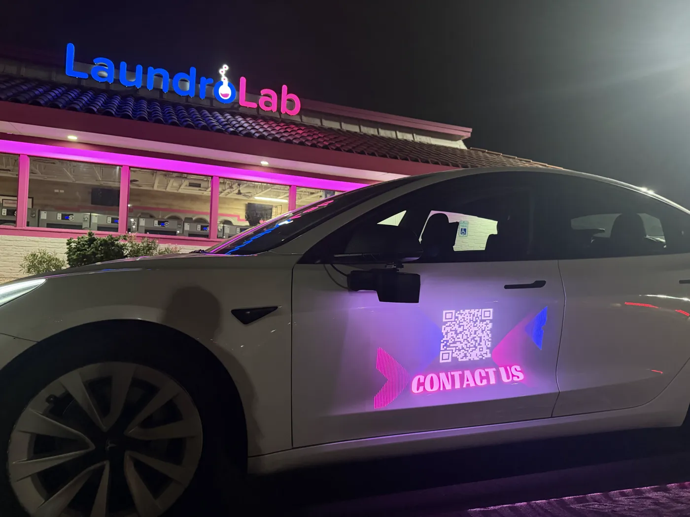 White Tesla at night with a scannable QR code and Contact Us message projected on its door
